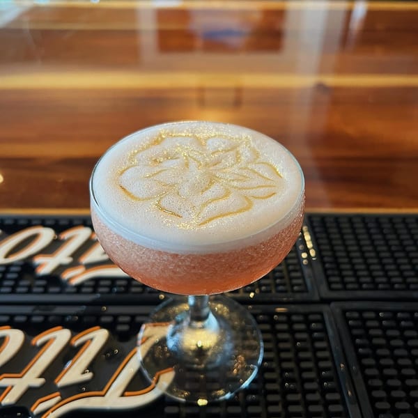 A Miyo's Restaurant cocktail with foamy top and a delicate flower design, on a bar mat.