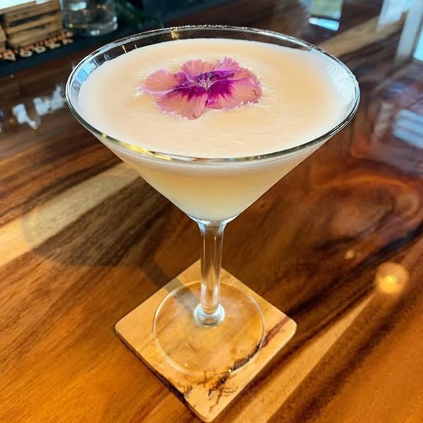 A creamy cocktail in a martini glass, garnished with a vibrant pink and purple flower, on a wooden coaster.