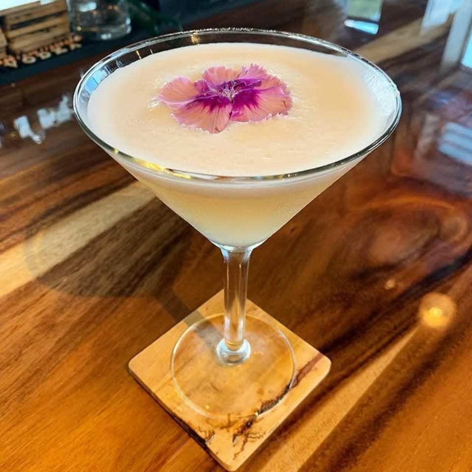 A creamy cocktail in a martini glass, garnished with a vibrant pink and purple flower, on a wooden coaster.