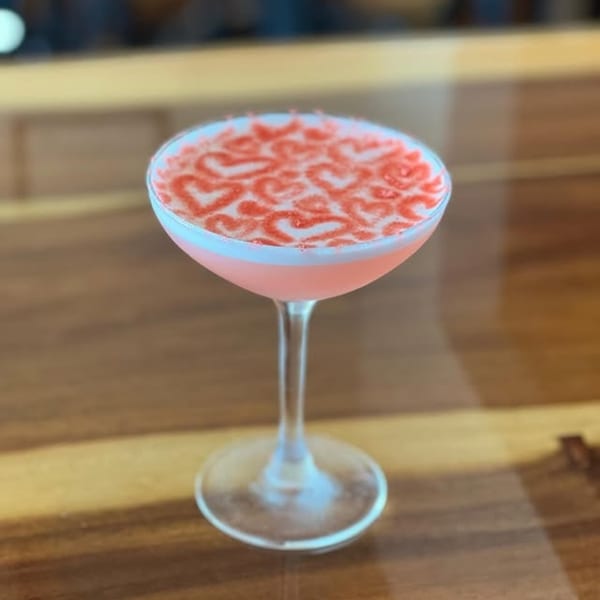Pink cocktail in a coupe glass, garnished with red heart designs on top, on a wooden table.