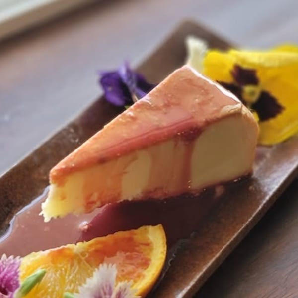Slice of Japanese cheesecake with red sauce, orange slices, and edible flowers on a brown plate.