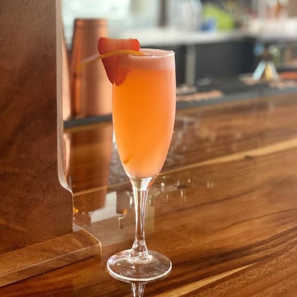 A pink cocktail in a champagne flute, garnished with a strawberry slice and lemon peel, on a wooden bar.