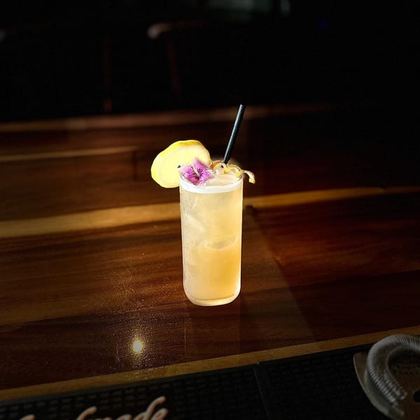 A tall, light-colored cocktail with a lemon slice, purple flower, and black straw on a wooden bar.