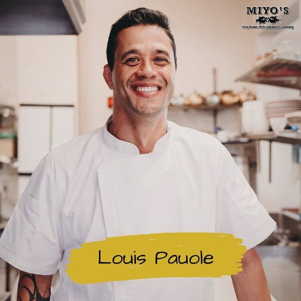 Smiling chef Louis Pauole in a white uniform, standing in a bright Miyo's Restaurant kitchen.