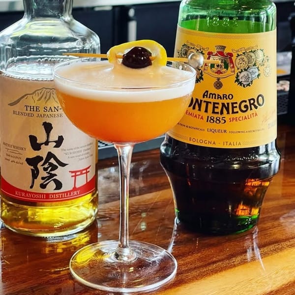 A Japanese whisky cocktail with a lemon and cherry garnish, next to bottles of whisky and Amaro Montenegro.